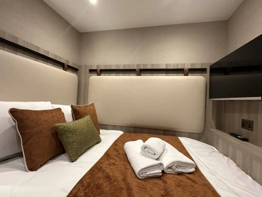 A neatly made bed with brown and green accent pillows, a brown throw, and two folded white towels. The room has light-colored walls, a padded headboard, and a wall-mounted flat-screen TV.