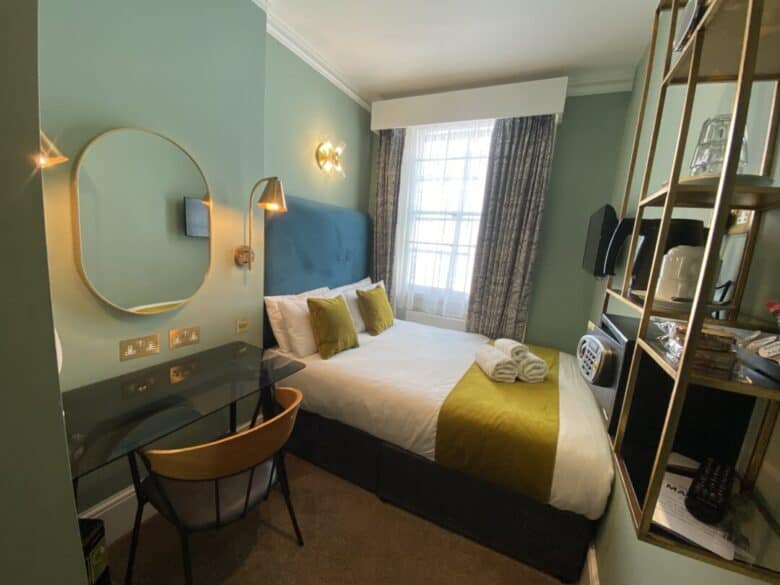 A small modern hotel room with a double bed, green accent pillows, a desk with a chair, a round mirror, wall lamps, shelves, and a window with patterned curtains letting in daylight. Towels are neatly placed on the bed.