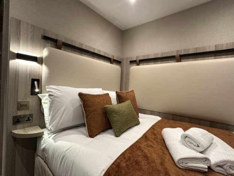 A neatly made bed with white sheets, brown and green cushions, and a brown blanket, with two folded white towels on top. The room has beige padded walls, wooden accents, and a wall light by the bed.