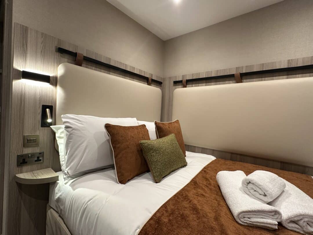 A neatly made bed with white sheets, brown and green cushions, and a brown blanket, with two folded white towels on top. The room has beige padded walls, wooden accents, and a wall light by the bed.