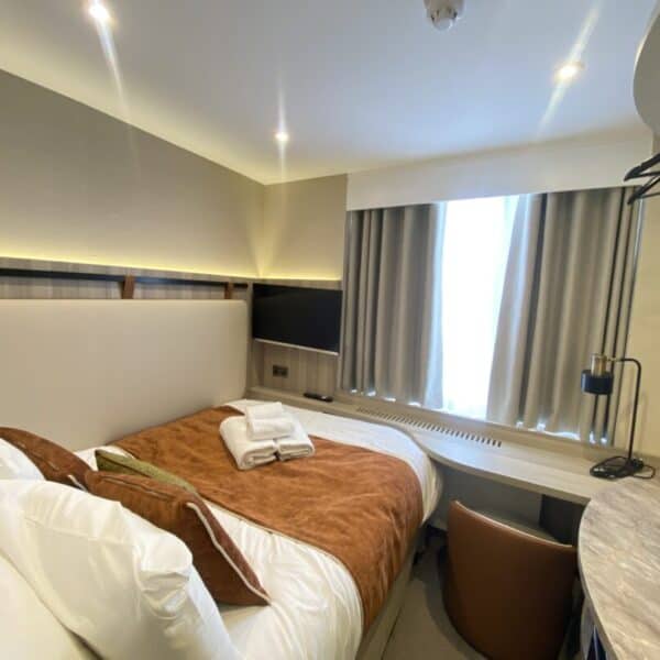 A compact, modern hotel room with a double bed, brown bedding, towels placed on top, a window with curtains, a desk and chair, a wall-mounted TV, and overhead lighting.