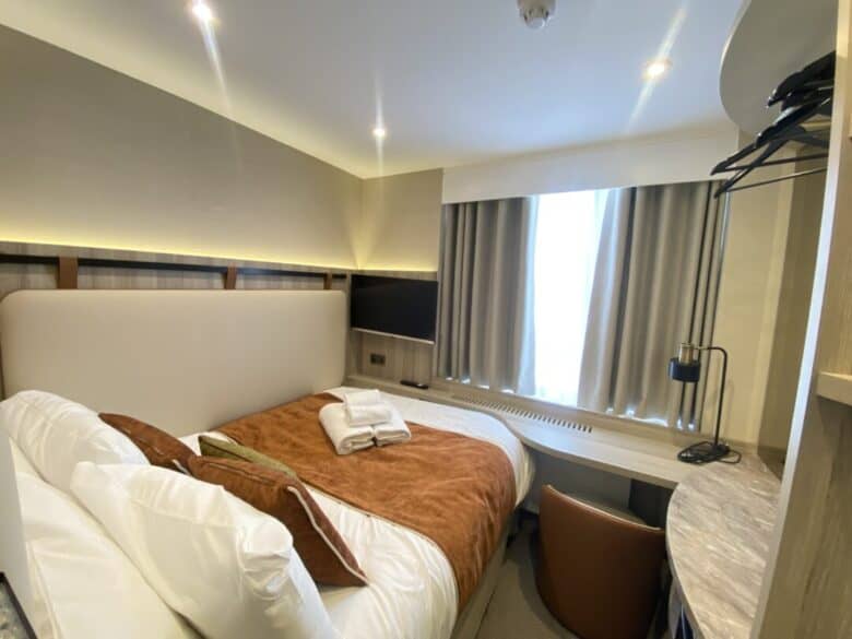 A compact, modern hotel room with a double bed, brown bedding, towels placed on top, a window with curtains, a desk and chair, a wall-mounted TV, and overhead lighting.