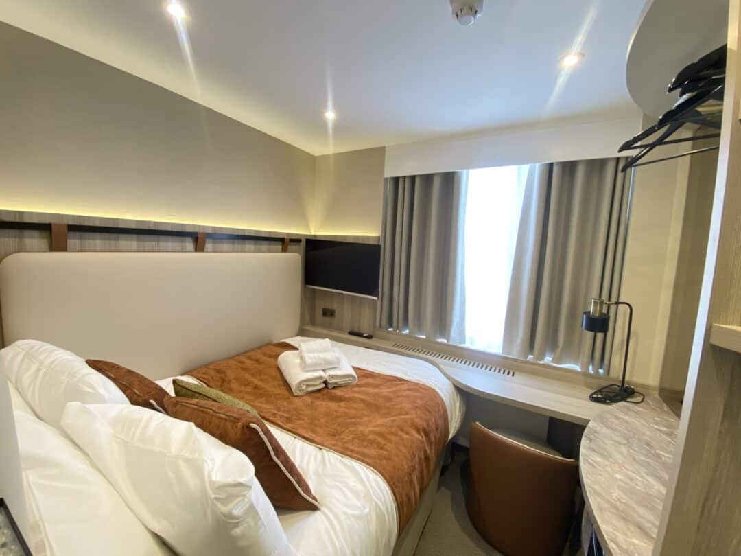 A compact, modern hotel room with a double bed, brown bedding, towels placed on top, a window with curtains, a desk and chair, a wall-mounted TV, and overhead lighting.