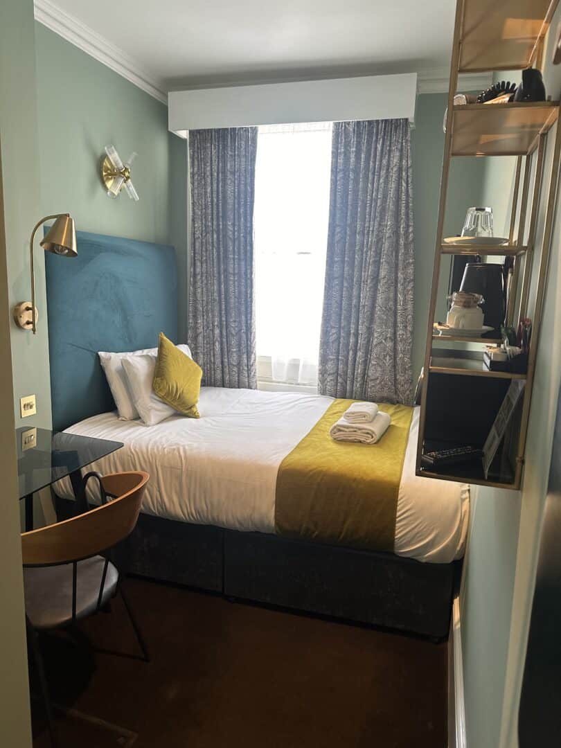 A small, neatly arranged hotel room with a single bed, yellow accents, a wall-mounted lamp, a chair at a glass desk, shelves with glasses and a kettle, and a window with patterned curtains letting in natural light.