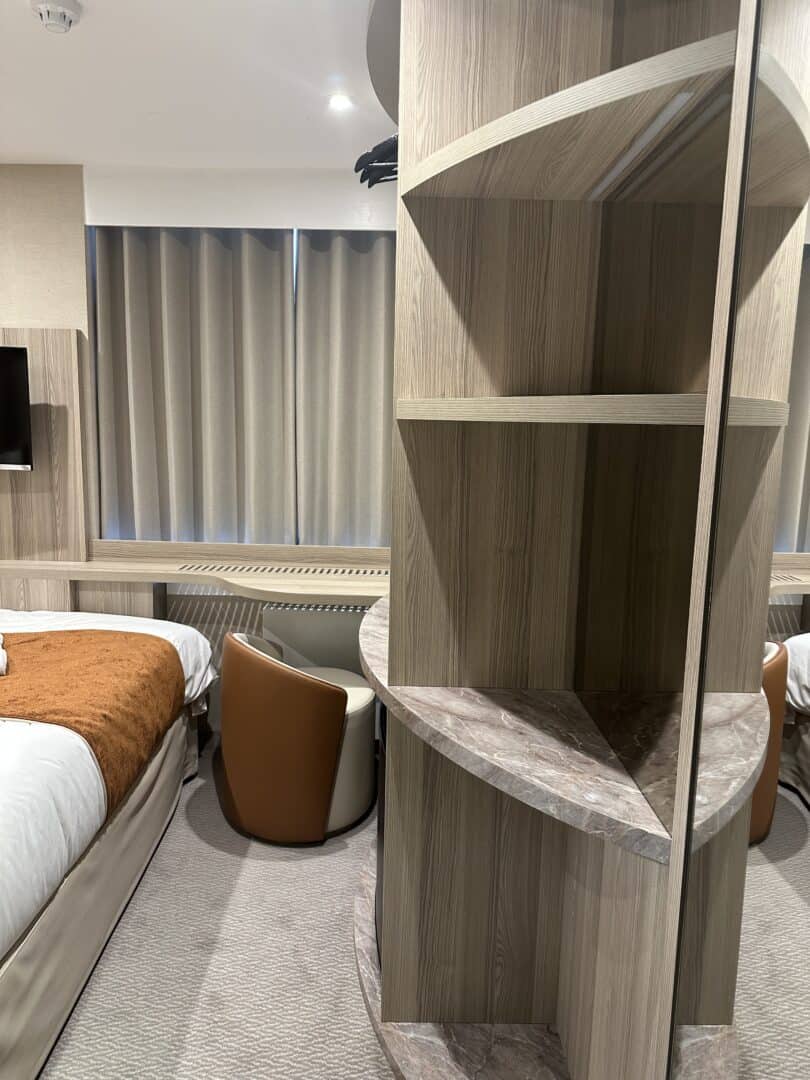 A modern hotel room with beige and brown tones features a bed with an orange throw, a round table with two brown chairs, and a curved wooden shelving unit dividing the space. Closed curtains cover the windows.