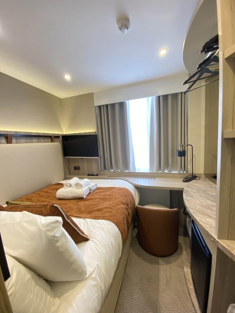 A compact hotel room with a neatly made bed, folded towels, a brown blanket, a small desk with a chair, wall-mounted TV, open closet, mini fridge, and a window with drawn curtains letting in daylight.