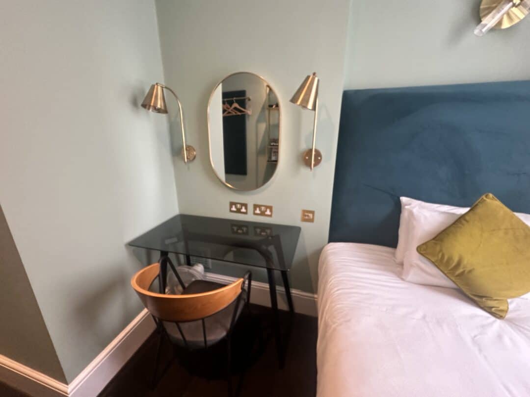A small hotel room corner with a glass-top desk, a wooden chair, a round mirror, two brass wall lamps, and part of a bed with a blue headboard and a green cushion.