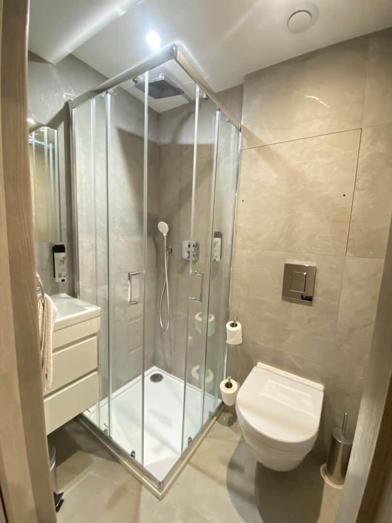 A modern, compact bathroom with beige tiles, a glass-enclosed corner shower, a wall-mounted toilet, a small white sink with drawers, and a toilet paper holder. Bright overhead lighting illuminates the space.