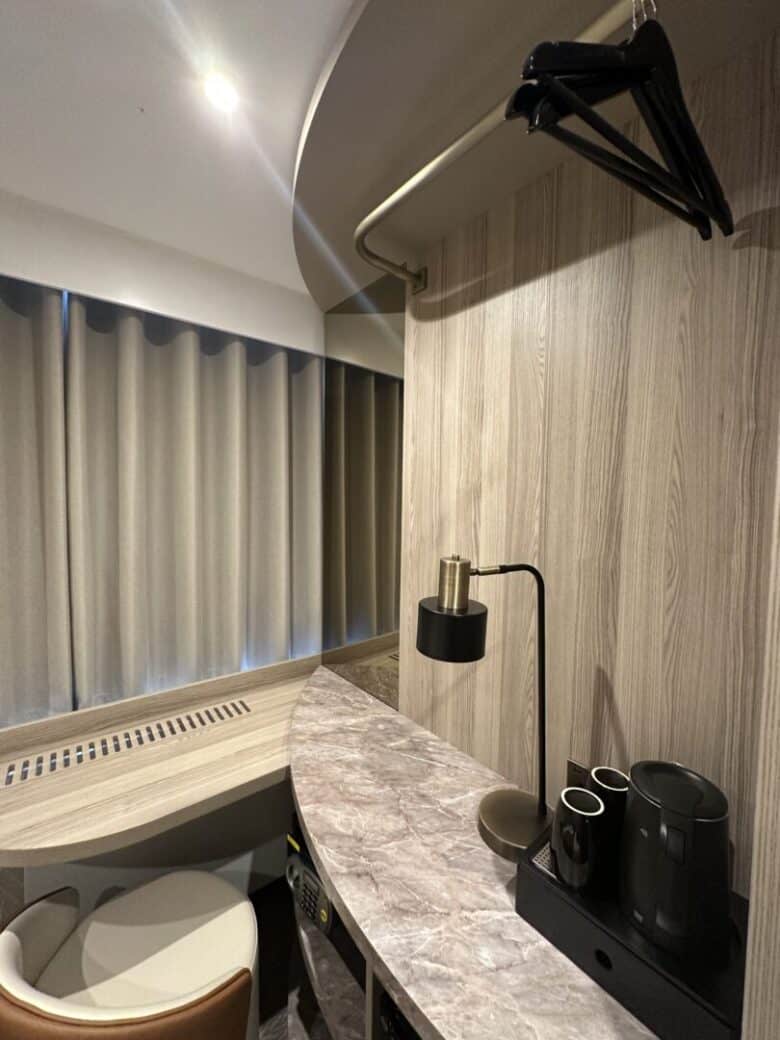 A small hotel room corner with a marble countertop, black electric kettle, two cups, a lamp, a chair, a curved wooden wall, hanging clothes hangers, and closed beige curtains over a window.