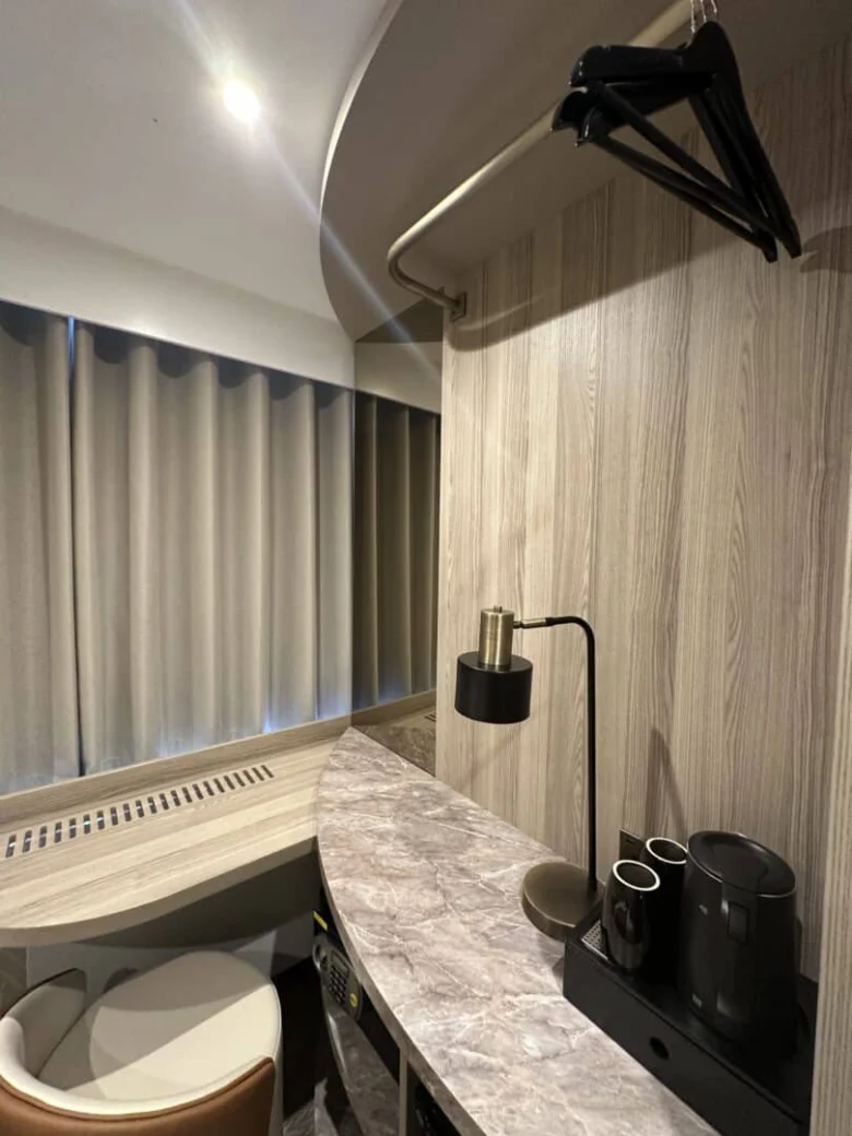 A small hotel room corner with a marble countertop, black electric kettle, two cups, a lamp, a chair, a curved wooden wall, hanging clothes hangers, and closed beige curtains over a window.