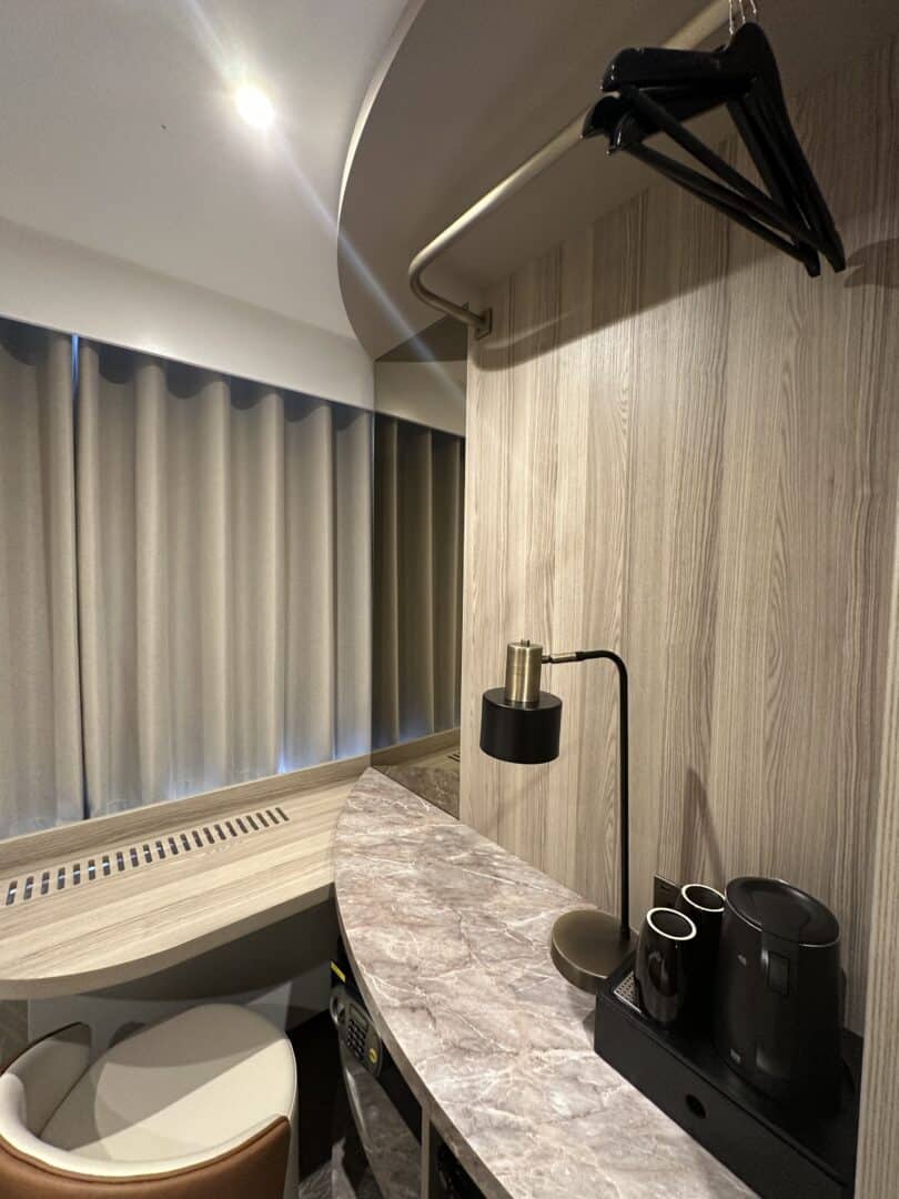 A small hotel room corner with a marble countertop, black electric kettle, two cups, a lamp, a chair, a curved wooden wall, hanging clothes hangers, and closed beige curtains over a window.