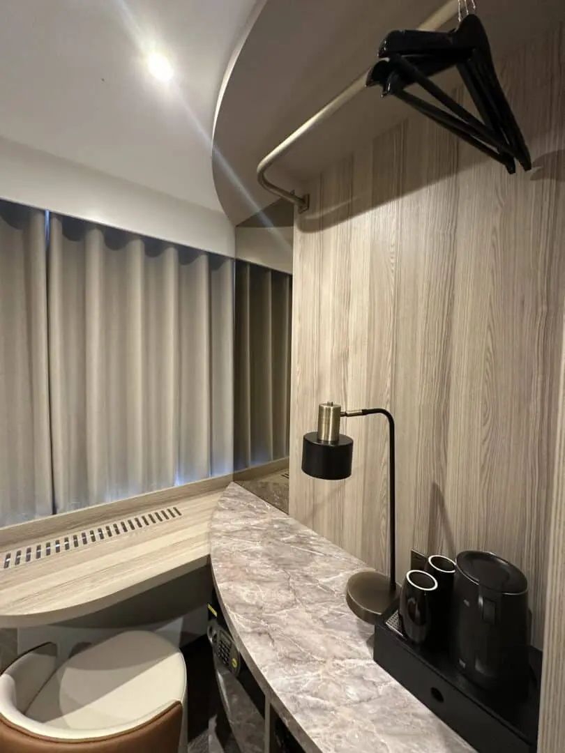 A small hotel room corner with a marble countertop, black electric kettle, two cups, a lamp, a chair, a curved wooden wall, hanging clothes hangers, and closed beige curtains over a window.