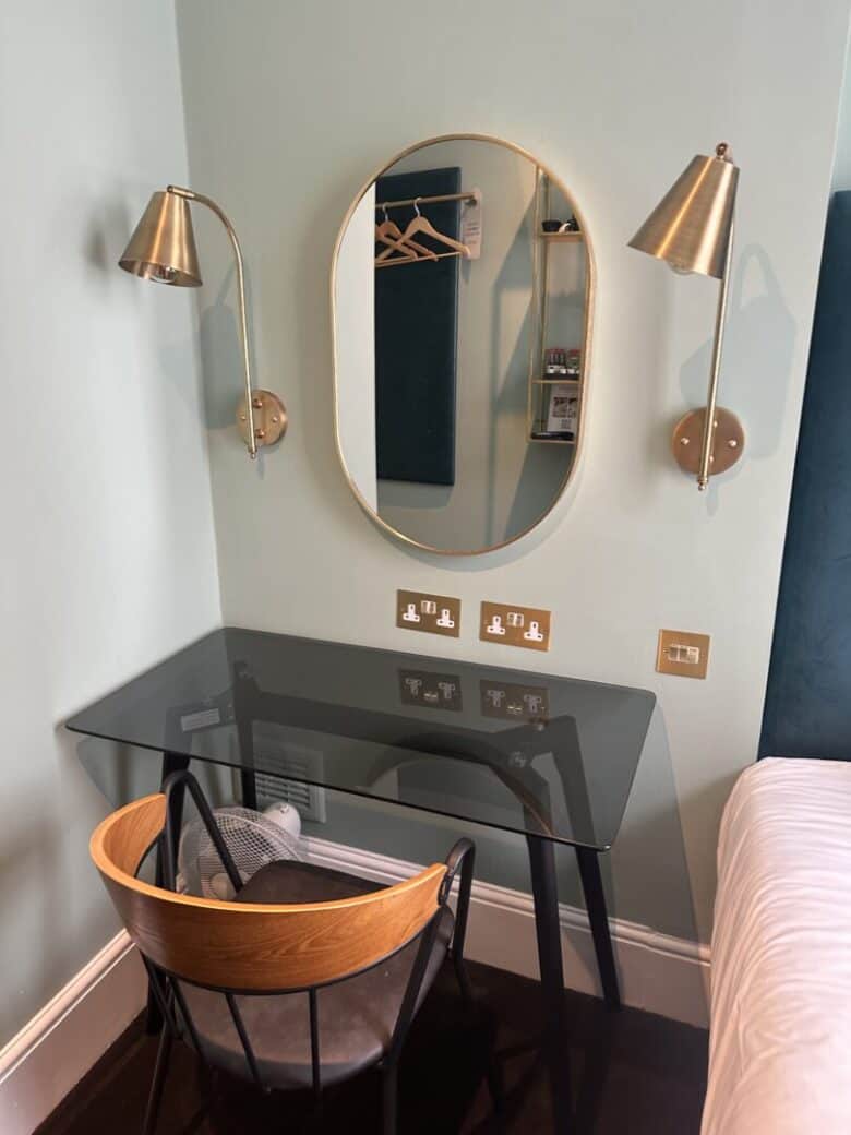 A modern workspace with a glass desk, wooden chair, oval wall mirror, and two brass wall-mounted lamps. The setup is next to a bed, with power outlets and light switches above the desk.