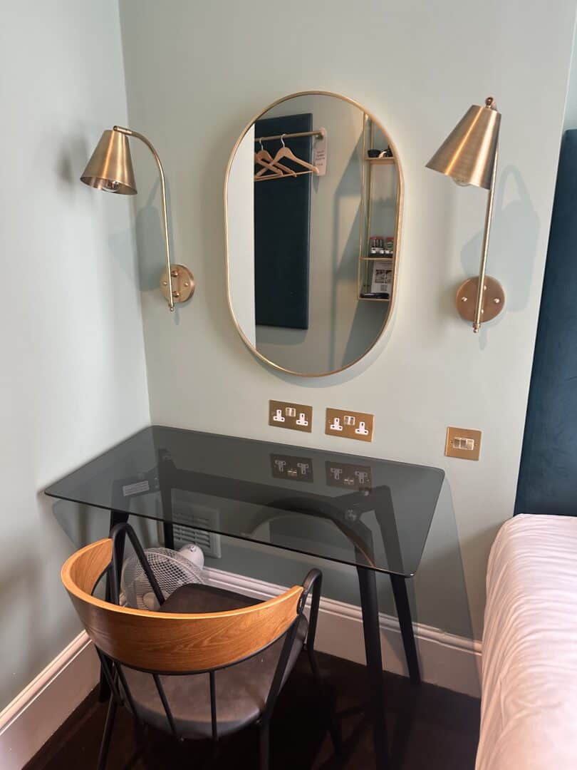 A modern workspace with a glass desk, wooden chair, oval wall mirror, and two brass wall-mounted lamps. The setup is next to a bed, with power outlets and light switches above the desk.