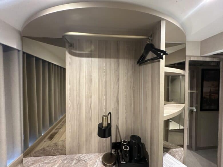 A modern hotel room corner with a wooden wardrobe, a hanging black hanger, a coffee machine, cups, a metal lamp, and a large round mirror reflecting part of the bathroom and hallway.