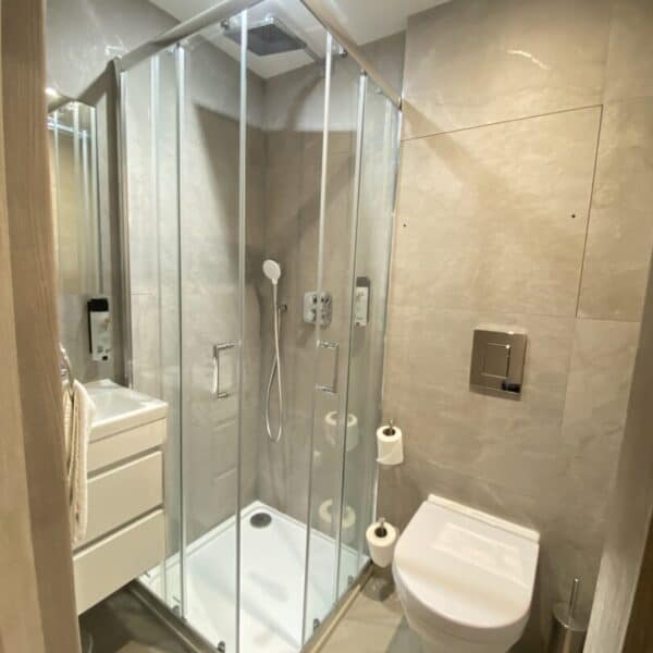 Modern bathroom with a glass shower enclosure, wall-mounted toilet, floating vanity with drawers, toilet paper holder, and neutral beige tiles on walls and floor. Bright and well-lit with a minimalist design.