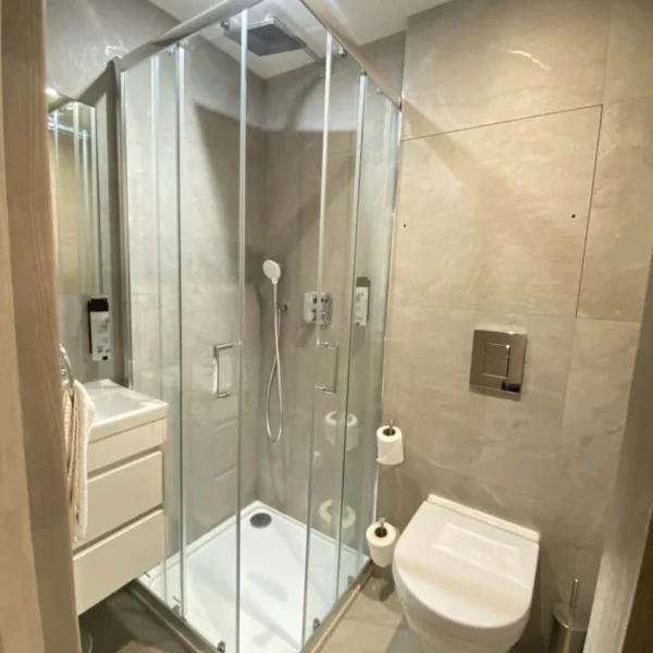 Modern bathroom with a glass shower enclosure, wall-mounted toilet, floating vanity with drawers, toilet paper holder, and neutral beige tiles on walls and floor. Bright and well-lit with a minimalist design.