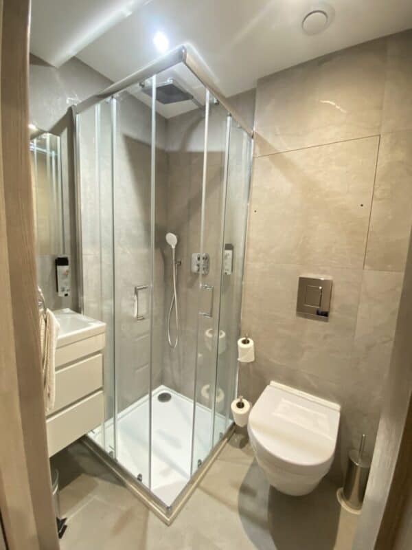 Modern bathroom with a glass shower enclosure, wall-mounted toilet, floating vanity with drawers, toilet paper holder, and neutral beige tiles on walls and floor. Bright and well-lit with a minimalist design.