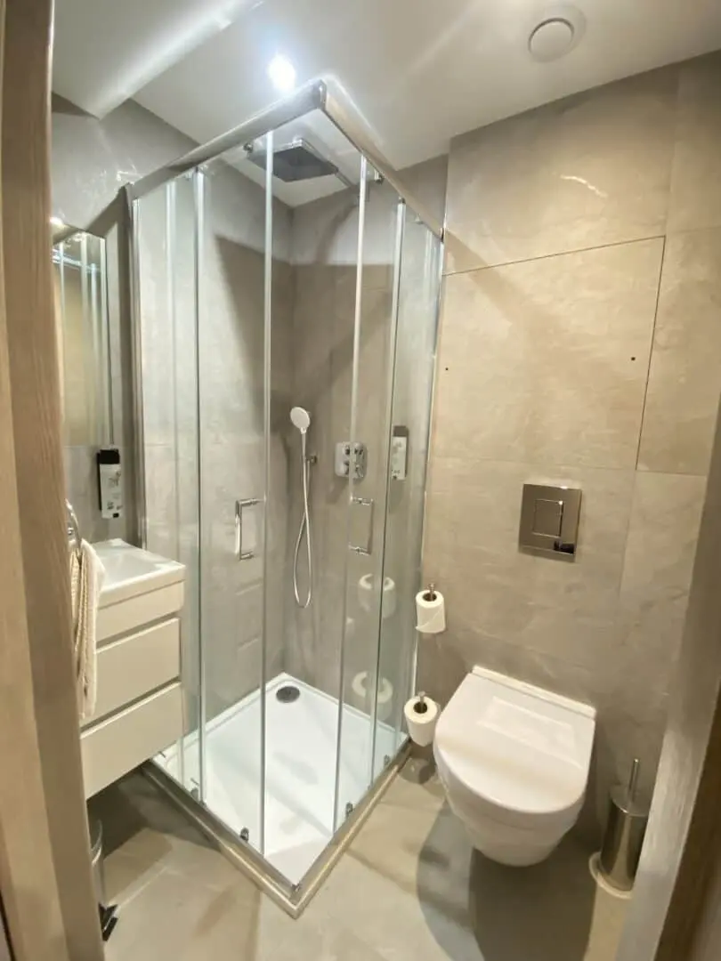 Modern bathroom with a glass shower enclosure, wall-mounted toilet, floating vanity with drawers, toilet paper holder, and neutral beige tiles on walls and floor. Bright and well-lit with a minimalist design.
