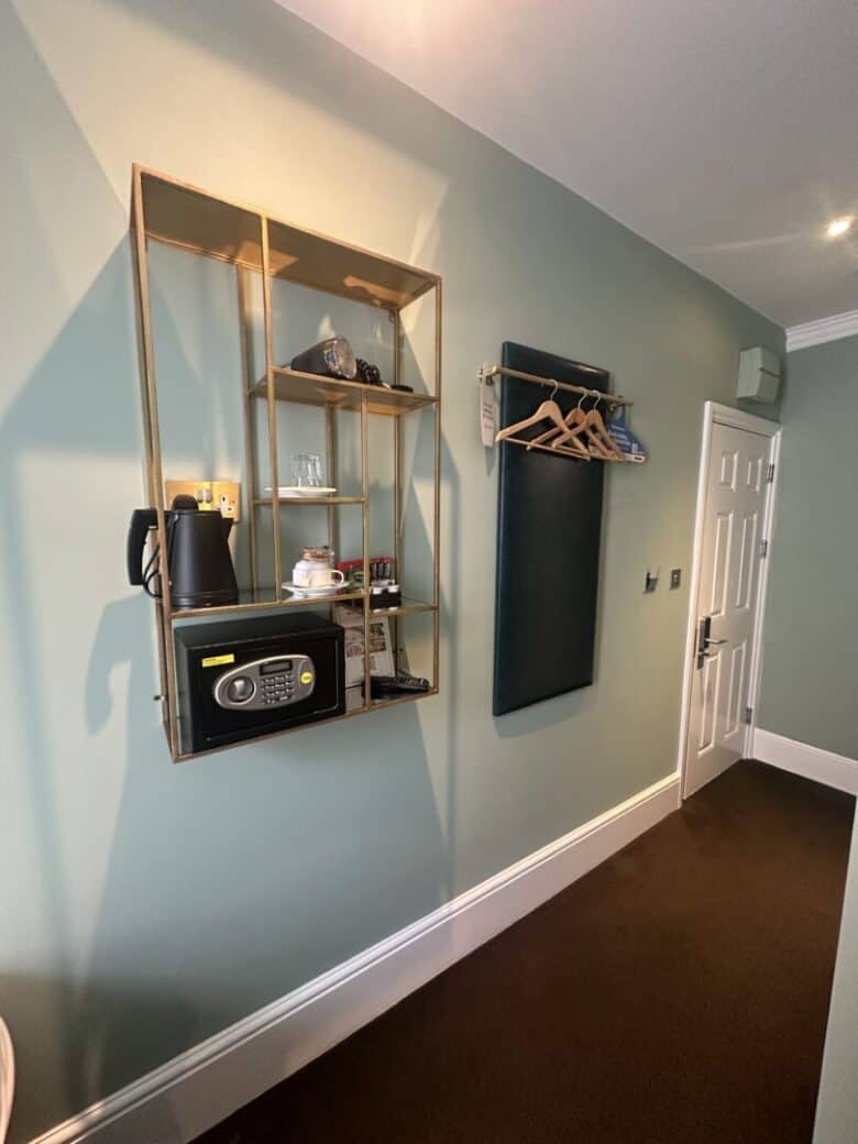 A hotel room entryway with a shelf holding a kettle, glasses, mugs, snacks, and a safe. To the right, a wall-mounted padded panel holds several wooden hangers near a white door.