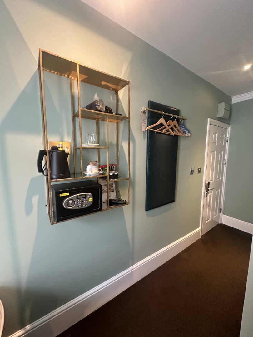 A hotel room entryway with a shelf holding a kettle, glasses, mugs, snacks, and a safe. To the right, a wall-mounted padded panel holds several wooden hangers near a white door.