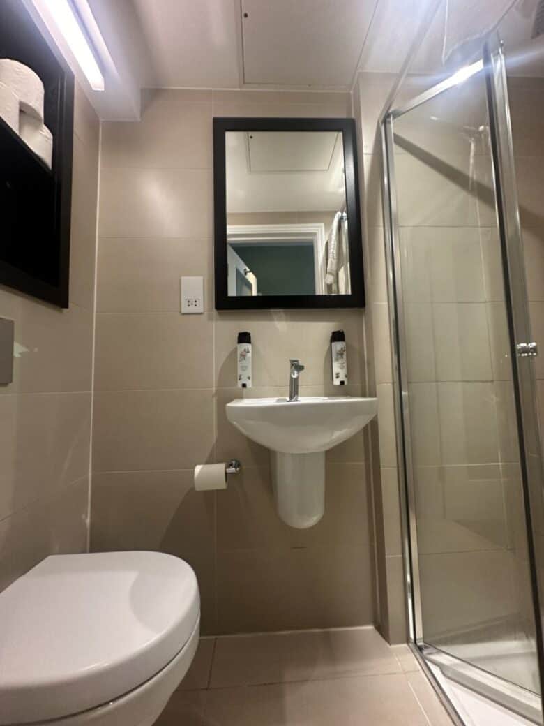 A modern bathroom with beige tiles, a wall-mounted sink beneath a black-framed mirror, two bottles on the sink, a glass shower enclosure on the right, and a toilet and toilet paper holder on the left.