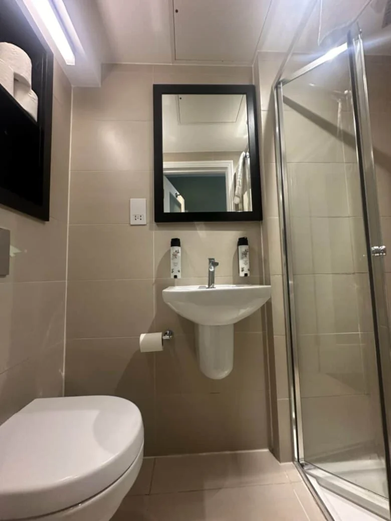 A modern bathroom with beige tiles, a wall-mounted sink beneath a black-framed mirror, two bottles on the sink, a glass shower enclosure on the right, and a toilet and toilet paper holder on the left.