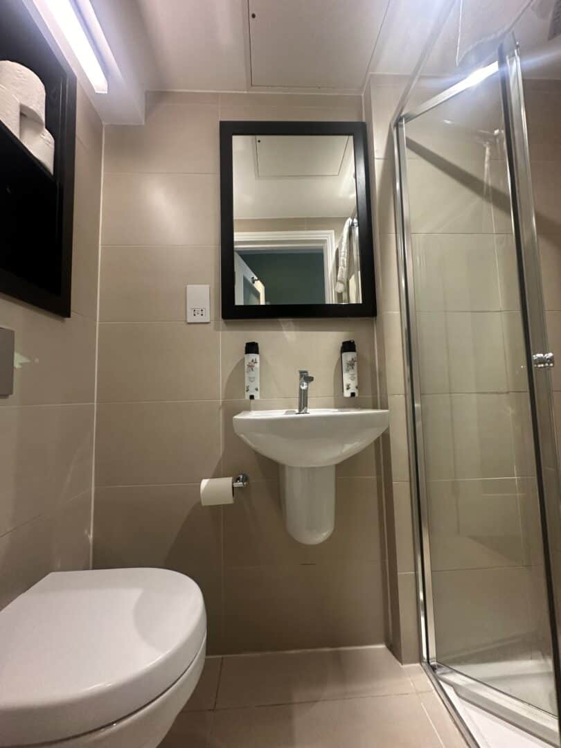 A modern bathroom with beige tiles, a wall-mounted sink beneath a black-framed mirror, two bottles on the sink, a glass shower enclosure on the right, and a toilet and toilet paper holder on the left.