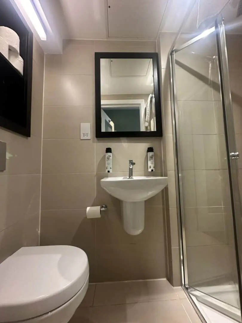 A modern bathroom with beige tiles, a wall-mounted sink beneath a black-framed mirror, two bottles on the sink, a glass shower enclosure on the right, and a toilet and toilet paper holder on the left.