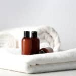Two brown plastic bottles with black caps sit on neatly folded white towels, with a rolled towel in the background, creating a clean and spa-like atmosphere.