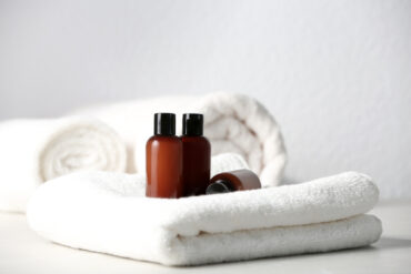 Two brown plastic bottles with black caps sit on neatly folded white towels, with a rolled towel in the background, creating a clean and spa-like atmosphere.