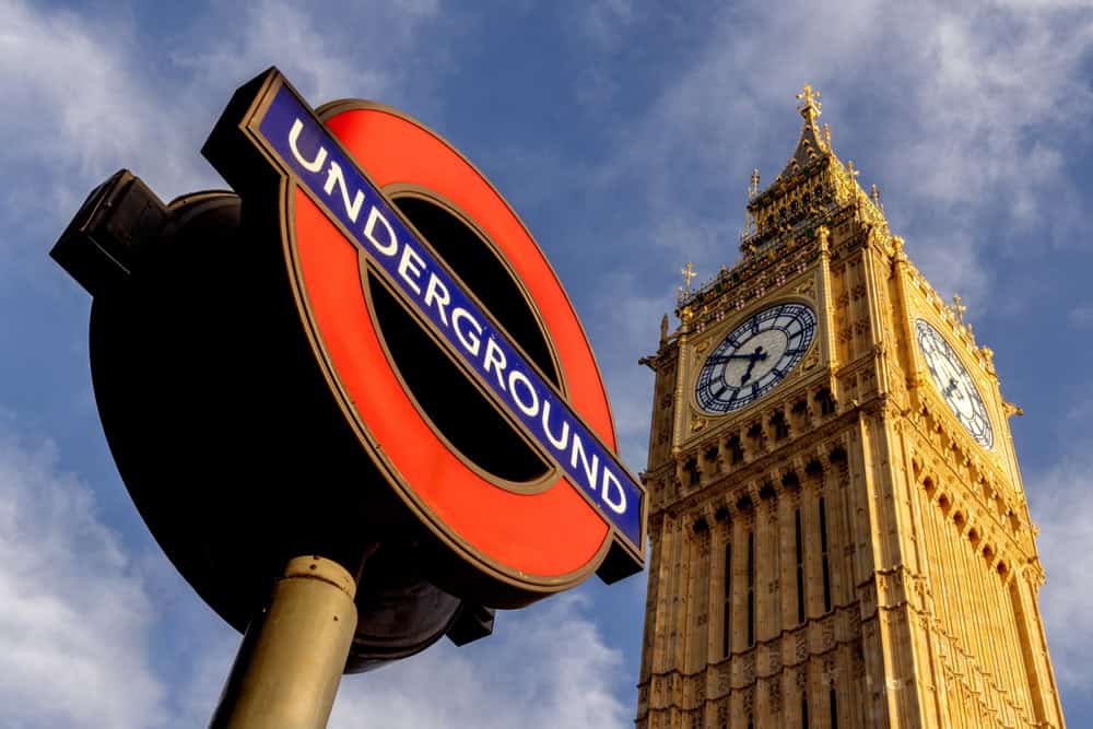 A London Underground sign in the foreground with the iconic Big Ben clock tower in the background, both under a blue sky with scattered clouds.
