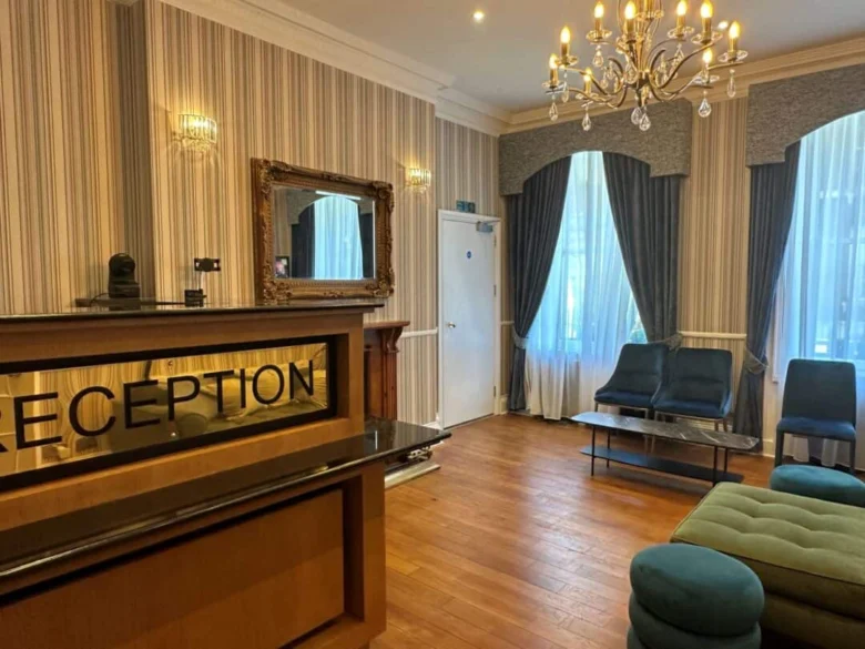 A hotel reception area with a wooden floor, reception desk, blue chairs and ottomans, large windows with blue curtains, a chandelier, and a wall mirror. The lighting is warm and elegant.