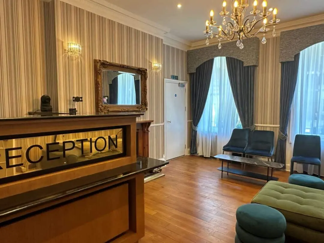 A hotel reception area with a wooden floor, reception desk, blue chairs and ottomans, large windows with blue curtains, a chandelier, and a wall mirror. The lighting is warm and elegant.