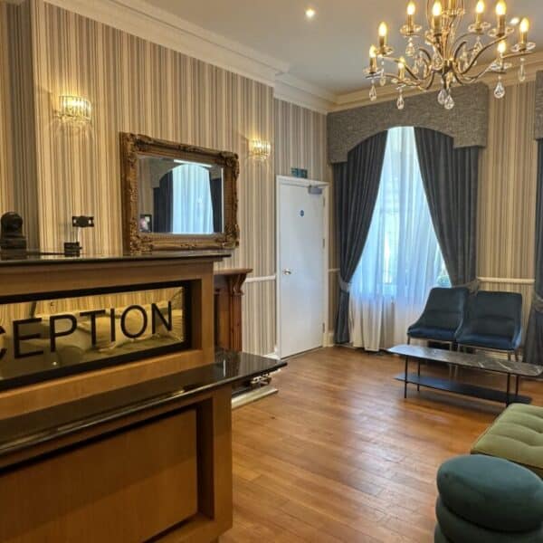Hotel reception area with a front desk, a large mirror, blue seating, wooden floors, striped wallpaper, and tall windows with curtains. A chandelier hangs from the ceiling, creating a welcoming atmosphere.