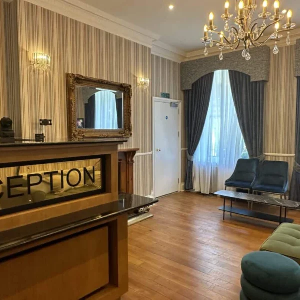 Hotel reception area with a front desk, a large mirror, blue seating, wooden floors, striped wallpaper, and tall windows with curtains. A chandelier hangs from the ceiling, creating a welcoming atmosphere.