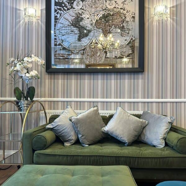 A green velvet sofa with four silver cushions sits in front of striped wallpaper. Above it hangs a framed world map. To the left, there is a gold and glass side table with a white orchid. A green ottoman is in front of the sofa.