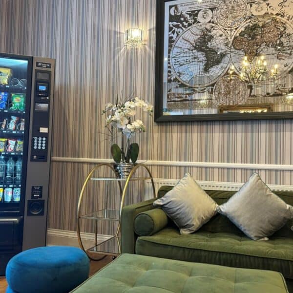 A vending machine stands next to a green sofa with silver cushions, a blue ottoman, a round table with a white orchid, and a large world map artwork on a striped wallpapered wall.