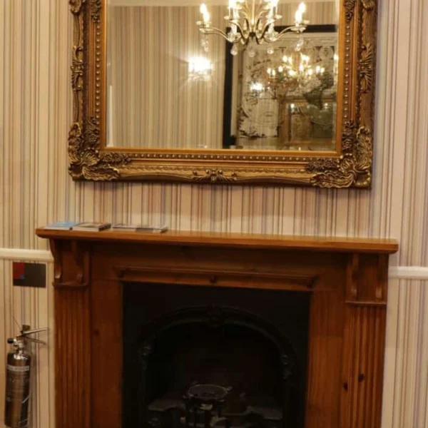 A wooden fireplace with a black cast iron insert is set against striped wallpaper. Above the mantel hangs a large ornate gold-framed mirror reflecting a chandelier and part of the room. A fire extinguisher is on the left.