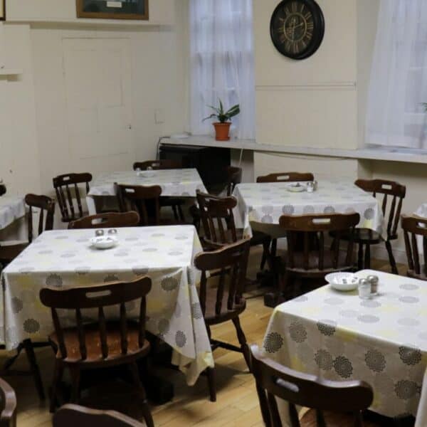 A small dining area with several wooden tables and chairs, each table covered with a patterned tablecloth and set with plates and shakers. Two potted plants sit on a windowsill beneath white curtains.