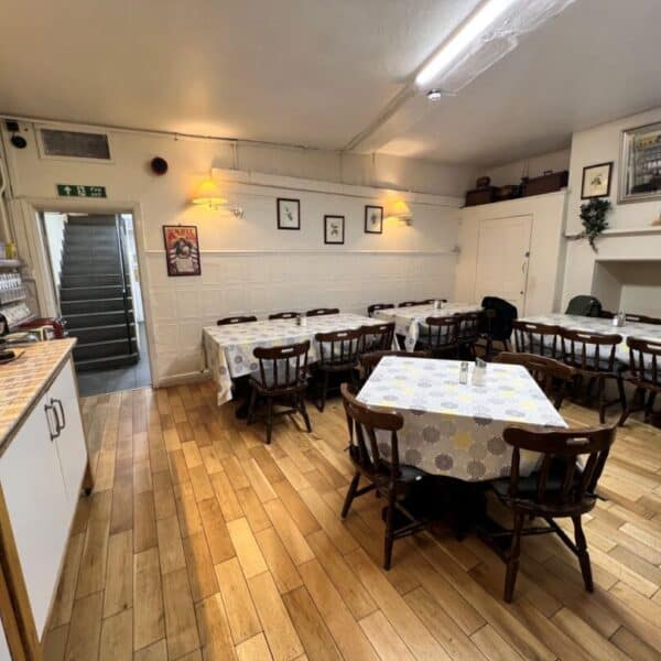 A cozy dining room with three tables covered in patterned tablecloths, wooden chairs, light wood flooring, wall art, and a counter with a coffee machine. Stairs and an exit are visible in the background.