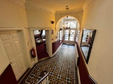 A hallway with patterned tile flooring, cream walls, and burgundy lower paneling leads to glass double doors. There are mirrors, a chandelier, and a fire extinguisher mounted on the wall. Doors open off the hallway.