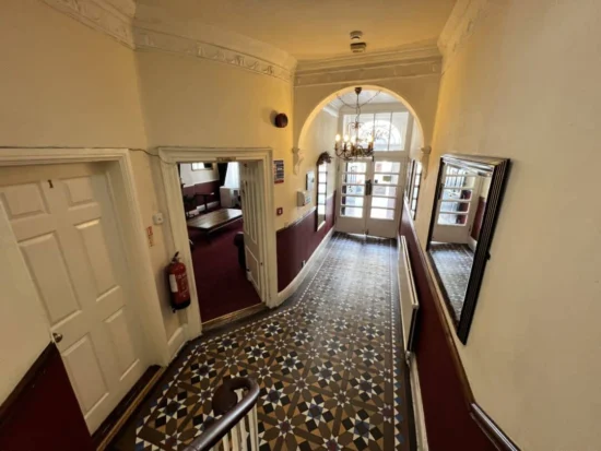 A hallway with patterned tile flooring, cream walls, and burgundy lower paneling leads to glass double doors. There are mirrors, a chandelier, and a fire extinguisher mounted on the wall. Doors open off the hallway.