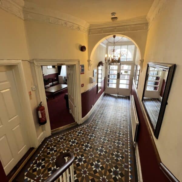 A hallway with patterned tile flooring, cream walls, and burgundy lower paneling leads to glass double doors. There are mirrors, a chandelier, and a fire extinguisher mounted on the wall. Doors open off the hallway.