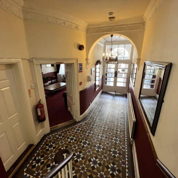 A hallway with patterned tile flooring, cream walls, and burgundy lower paneling leads to glass double doors. There are mirrors, a chandelier, and a fire extinguisher mounted on the wall. Doors open off the hallway.