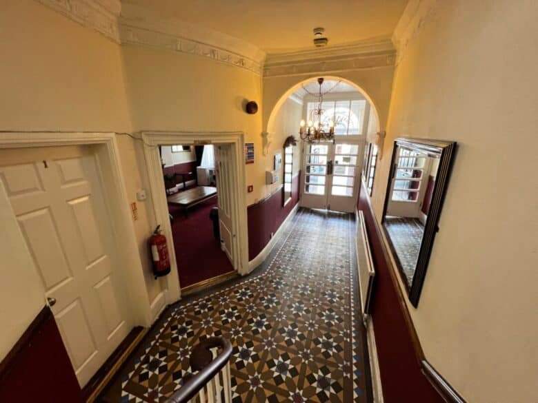 A hallway with patterned tile flooring, cream walls, and burgundy lower paneling leads to glass double doors. There are mirrors, a chandelier, and a fire extinguisher mounted on the wall. Doors open off the hallway.