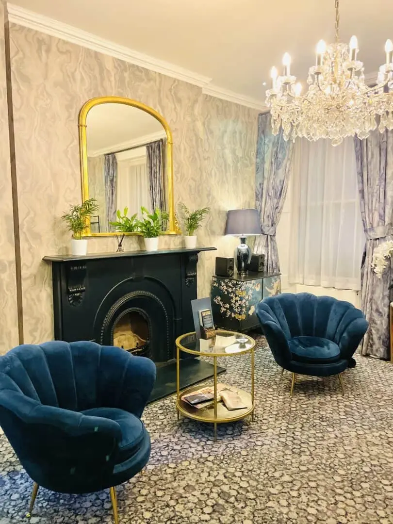 Elegant living room with two blue velvet chairs, a round glass coffee table, ornate black fireplace with a large gold-framed mirror, crystal chandelier, floral curtains, and a patterned carpet.