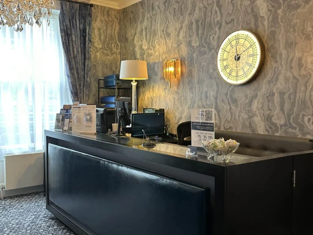 A hotel reception desk with a lamp, brochures, a sign with a QR code, and a bowl of flowers. Behind the desk is a large decorative clock on a patterned wall and a window with sheer curtains.