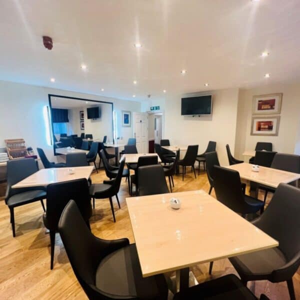 A well-lit dining area with several light wood tables and black chairs, wooden flooring, a wall-mounted TV, framed pictures, and shelves with dishes in the corner. The room has a modern and clean appearance.