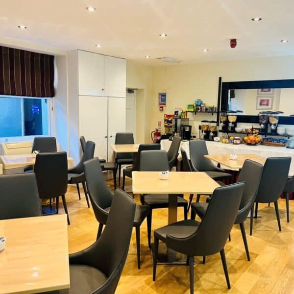 A bright, modern dining area with wooden floors, several tables and black chairs. A buffet with fruit, drinks, and breakfast items is set up along one wall. The room has large windows and light-colored walls.